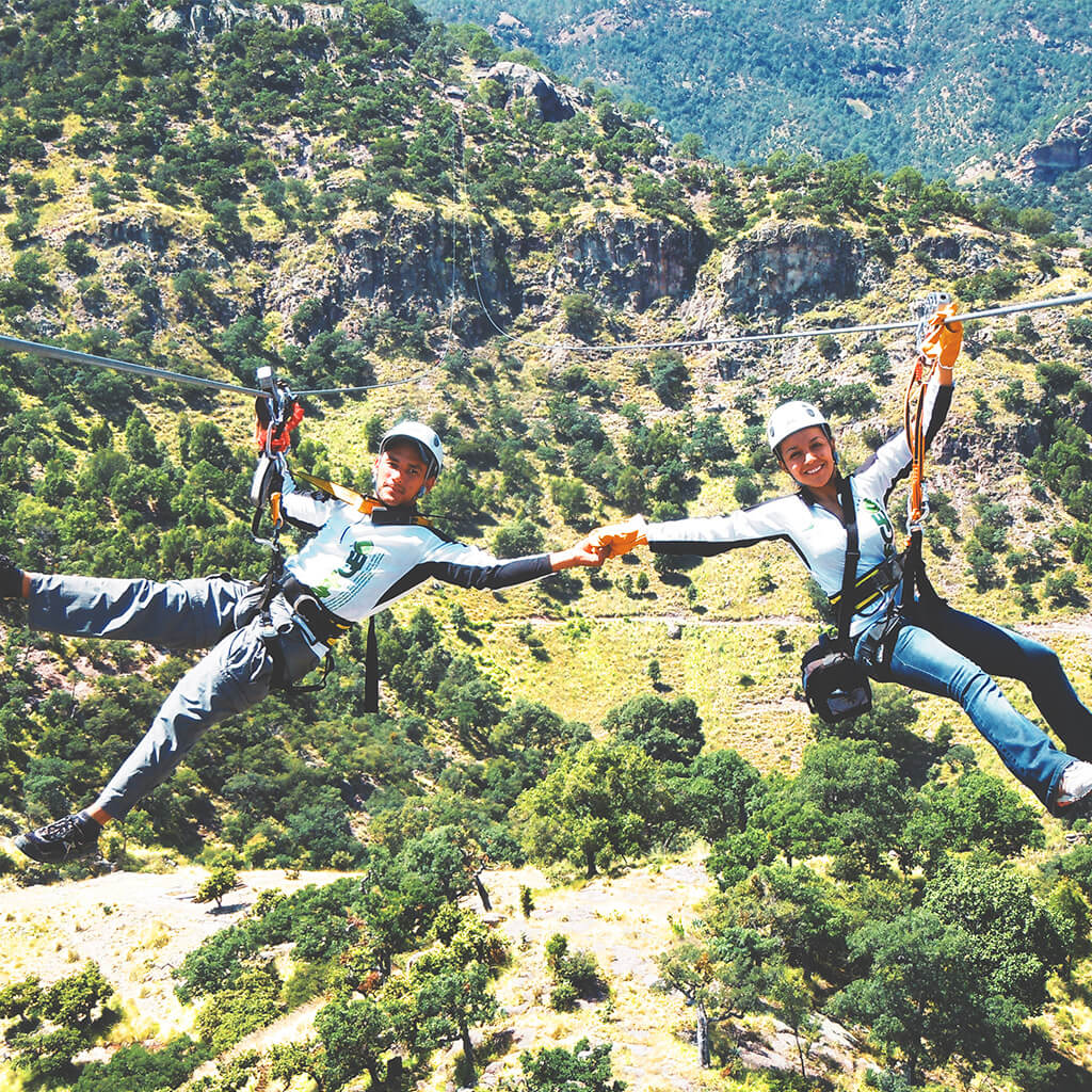 Ecoprojects. Parque de Aventura Barrancas del Cobre. Dos personas compartiendo una tirolesa sobre una montaña llena de árboles.