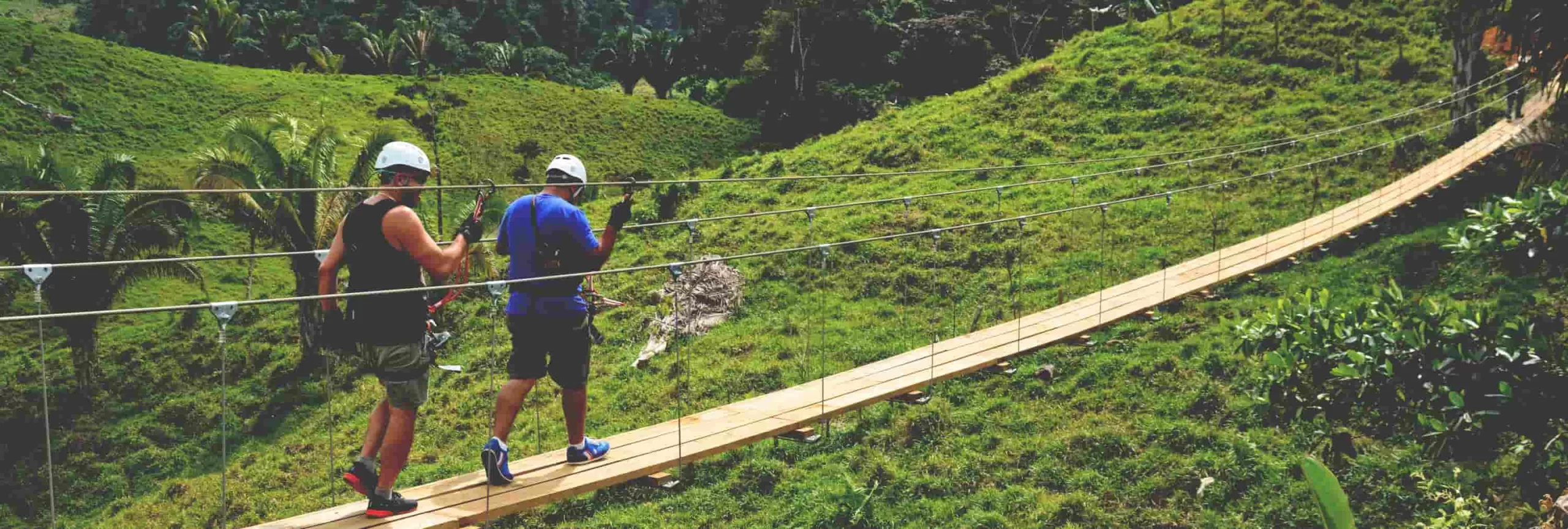 Ecoproparks. Dos personas equipadas con protección caminando de manera segura sobre un puente de madera, disfrutando de una aventura en medio de una montaña verde y exuberante con Ecoproparks.